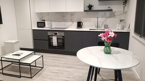 Een moderne keuken met witte kasten, een magnetron, een oven en een koffiezetapparaat. Er staat een eettafel met een bloemenvaas en een kleine bijzettafel.
