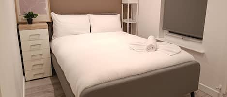 A neatly made bed with a rolled towel, a bedside table with drawers, a lamp, and a framed artwork on the wall.