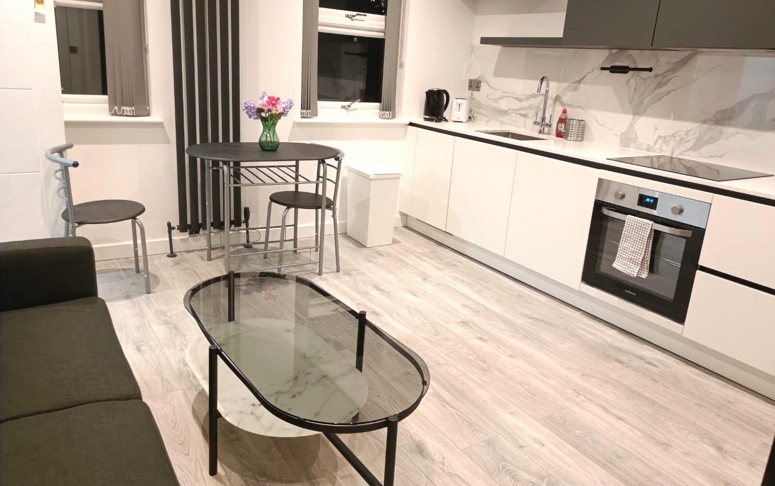 Une cuisine moderne avec une table à manger au plateau en verre, un four encastré et une fenêtre avec des rideaux.