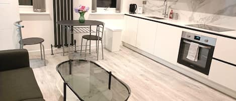 A modern kitchen with a glass-top dining table, a built-in oven, and a window with curtains.
