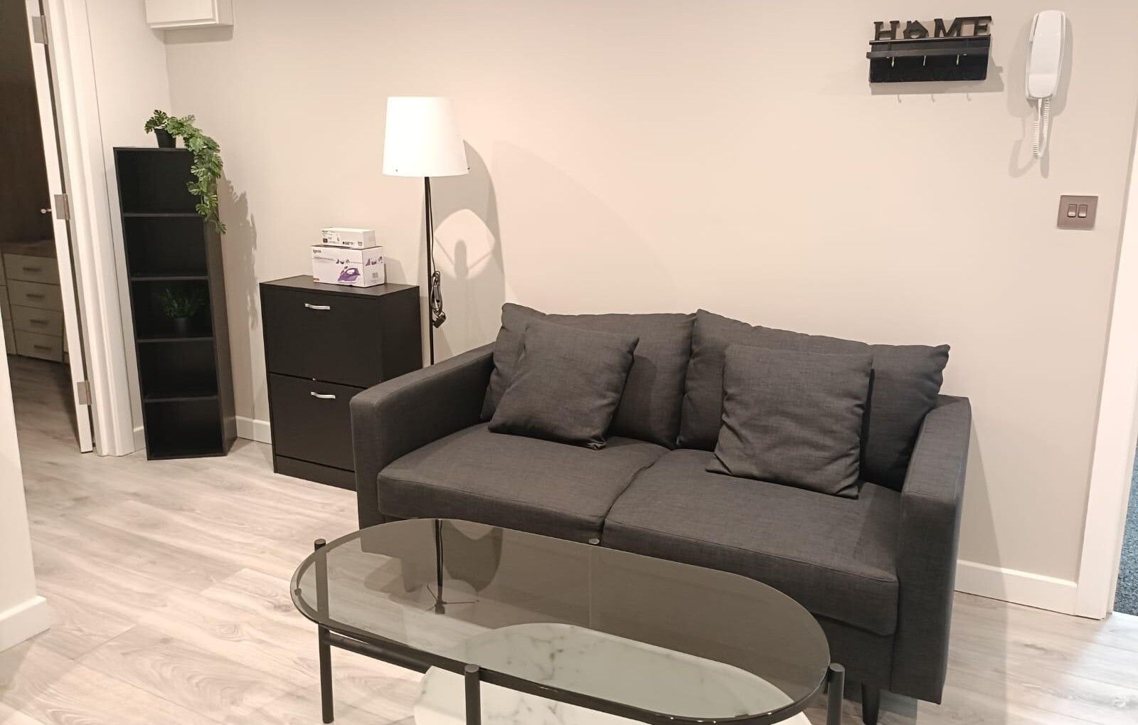 Un salon moderne avec un canapé gris, une table basse en verre, une table d’appoint noire avec une lampe, et un panneau décoratif portant l’inscription « HOME » au mur.