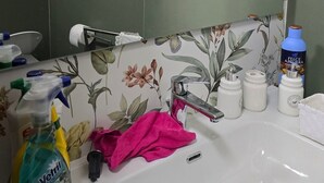 Een badkamer met een wastafel, spiegel en groene tegels. Er staan schoonmaakmiddelen en een handdoek op het aanrecht.