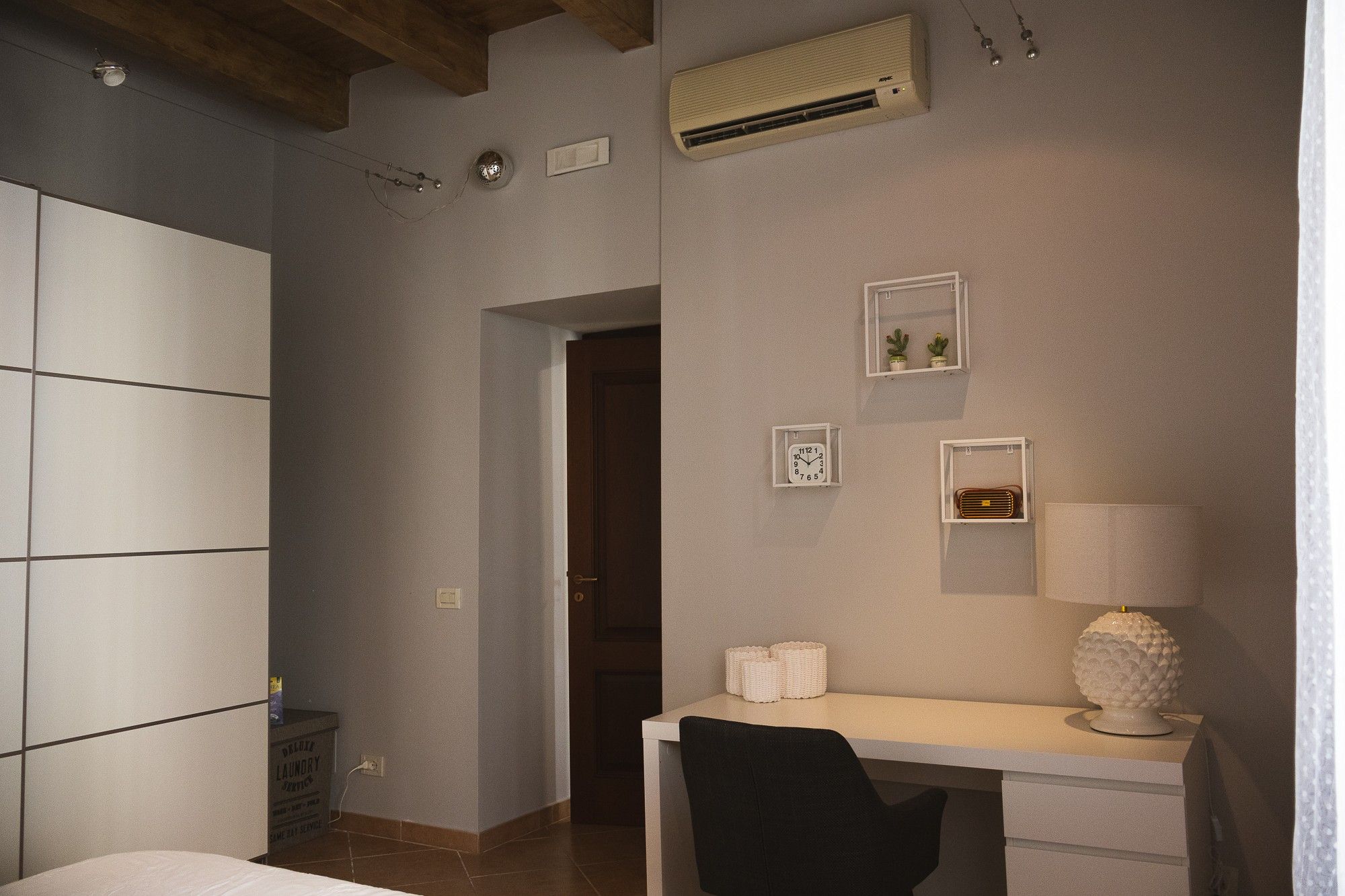 Un dormitorio con cama, escritorio, silla, aire acondicionado de pared y puerta que da a otra habitación.