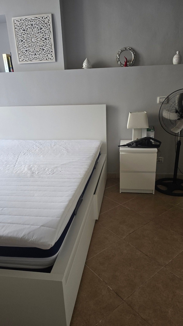 Un dormitorio con cama, mesita de noche, lámpara y ventilador de pie.