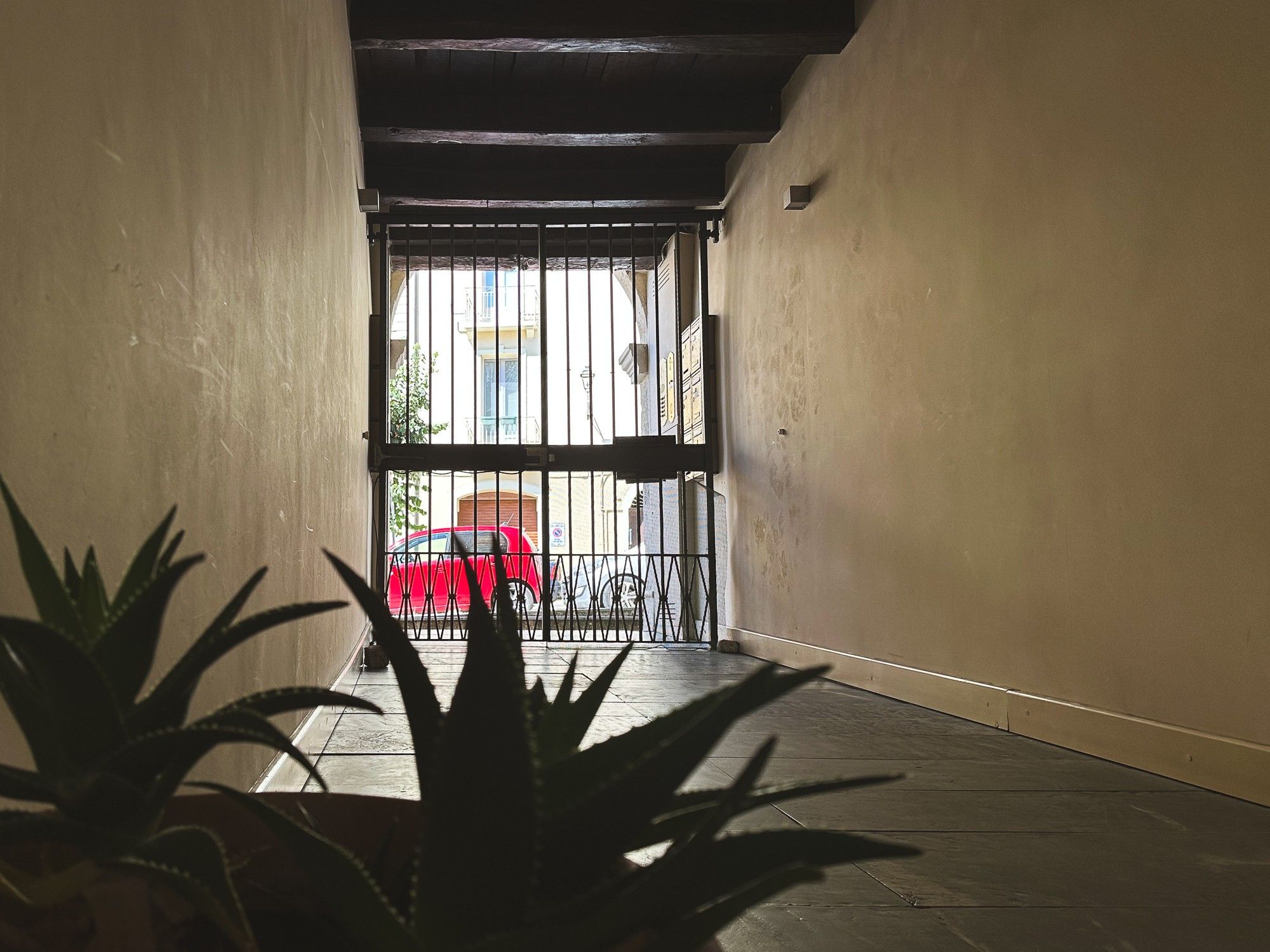 A corridor with a metal gate leading to an outdoor area with a red chair and a bicycle.