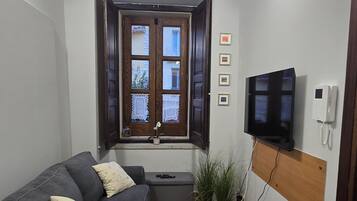 Un espacio habitable compacto con un sofá, un televisor montado en la pared, techo de madera y una ventana con cortinas de encaje.