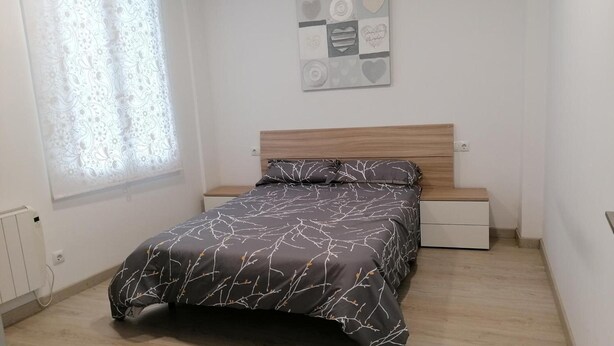 Un dormitori amb un llit, una tauleta de nit, un moble amb calaixos, una finestra amb cortines i una decoració penjada a la paret.
