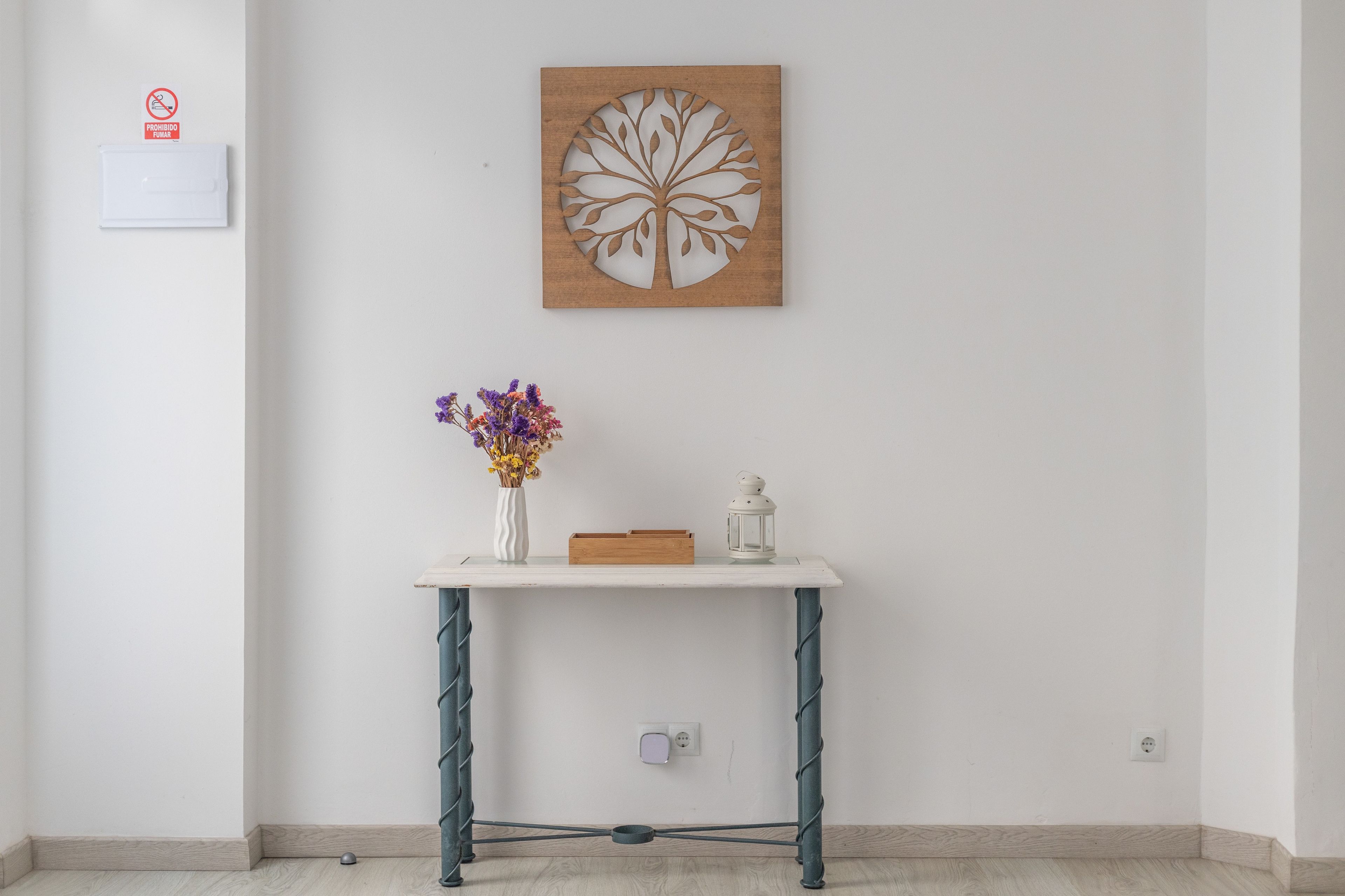 Una habitación con pared blanca, una obra de arte de madera, un florero blanco con flores, un farol blanco y un pequeño cofre de madera sobre una mesa blanca.