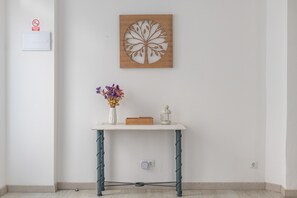 Habitació amb una paret blanca, una obra d'art de fusta, un vas blanc amb flors, un fanal blanc i una petita caixa de fusta sobre una taula blanca.
