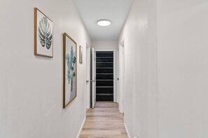 A hallway with framed artwork on the walls, a wooden floor, and a staircase leading to a door. - Poconos Home | Hot Tub, Cold Plunge & Fire Pit (Long Pond)