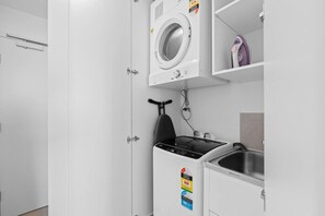 A compact laundry area with a washing machine, a dryer, a small ironing board, and a sink. - Stylish 1BR in the Heart of Broadbeach (Broadbeach)