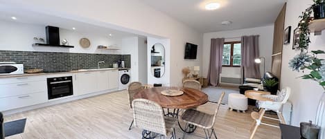 A modern kitchen with white cabinets, a microwave, and a washing machine. There is a dining area with a wooden table and wicker chairs.