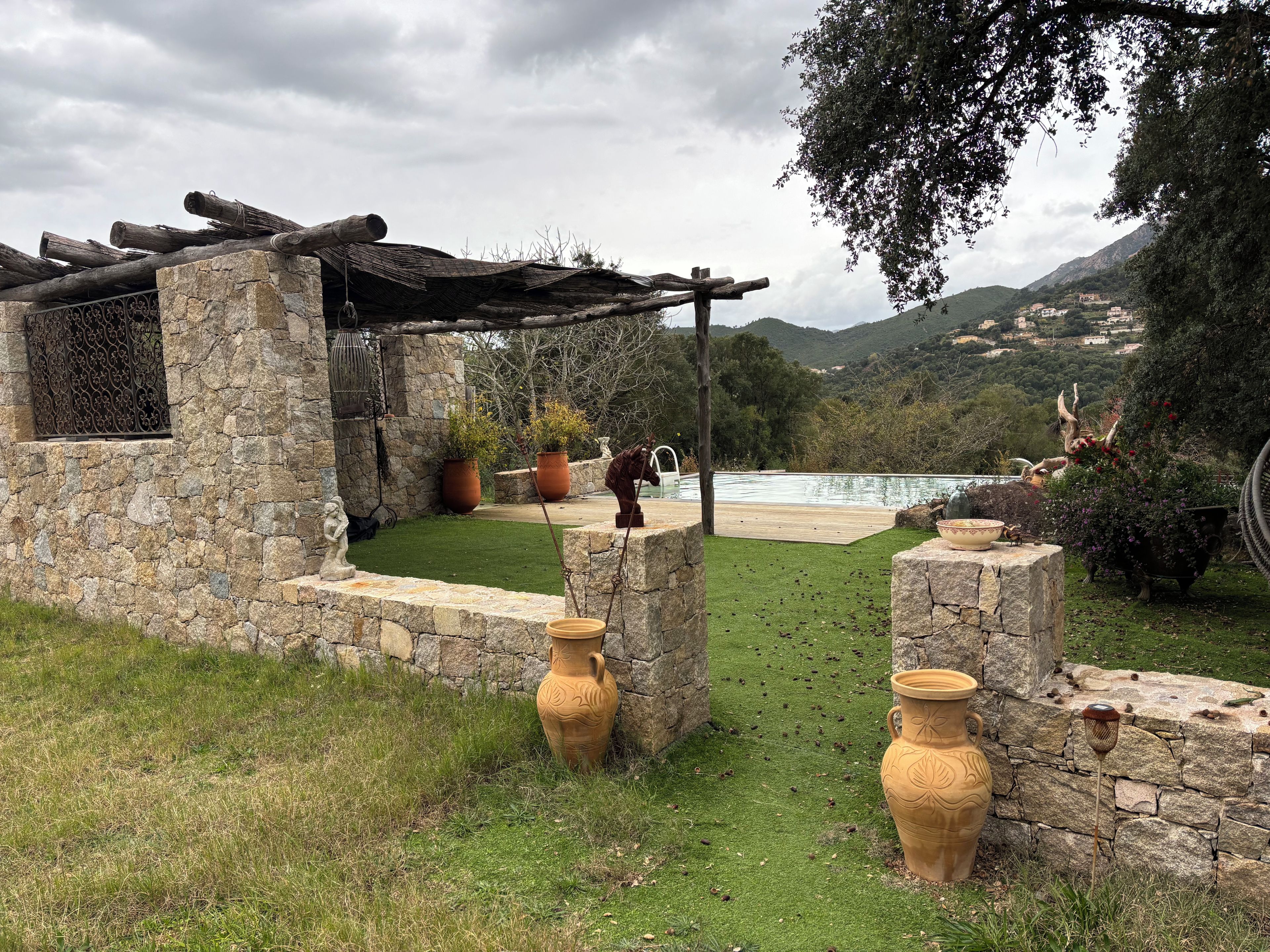 Une construction en pierre avec une piscine, entourée de verdure et adossée à un paysage montagneux.