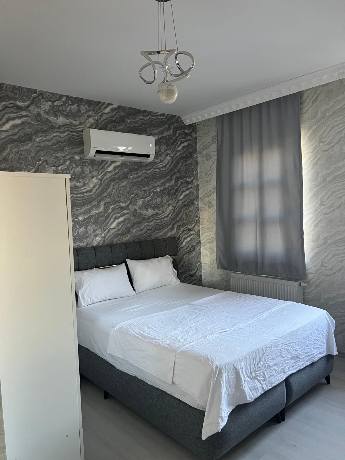 Quarto moderno com cama, paredes cinzentas, janela com cortinas e ar condicionado.