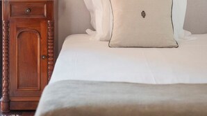 Una cama bien tendida con sábanas blancas y una única almohada decorativa. A la izquierda de la cama, una mesita de noche con una lámpara.