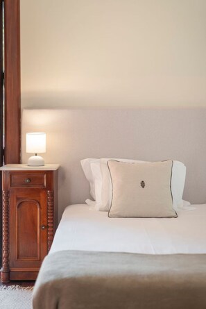 A neatly made bed with white linens and a single decorative pillow. A wooden nightstand with a lamp is positioned to the left of the bed. - Vrbo Property (Santana)