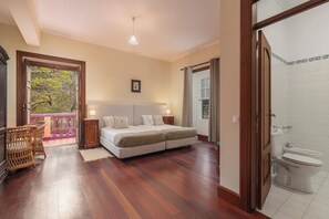 Ein Schlafzimmer mit Doppelbett, Nachttisch, Korbsessel, einem Balkon mit Blick auf Bäume und einem Badezimmer mit Toilette und Waschbecken.
