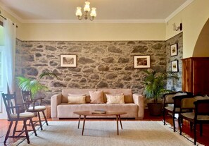 A living room with a stone wall, a beige sofa, wooden chairs, a coffee table, and a chandelier. - Vrbo Property (Santana)