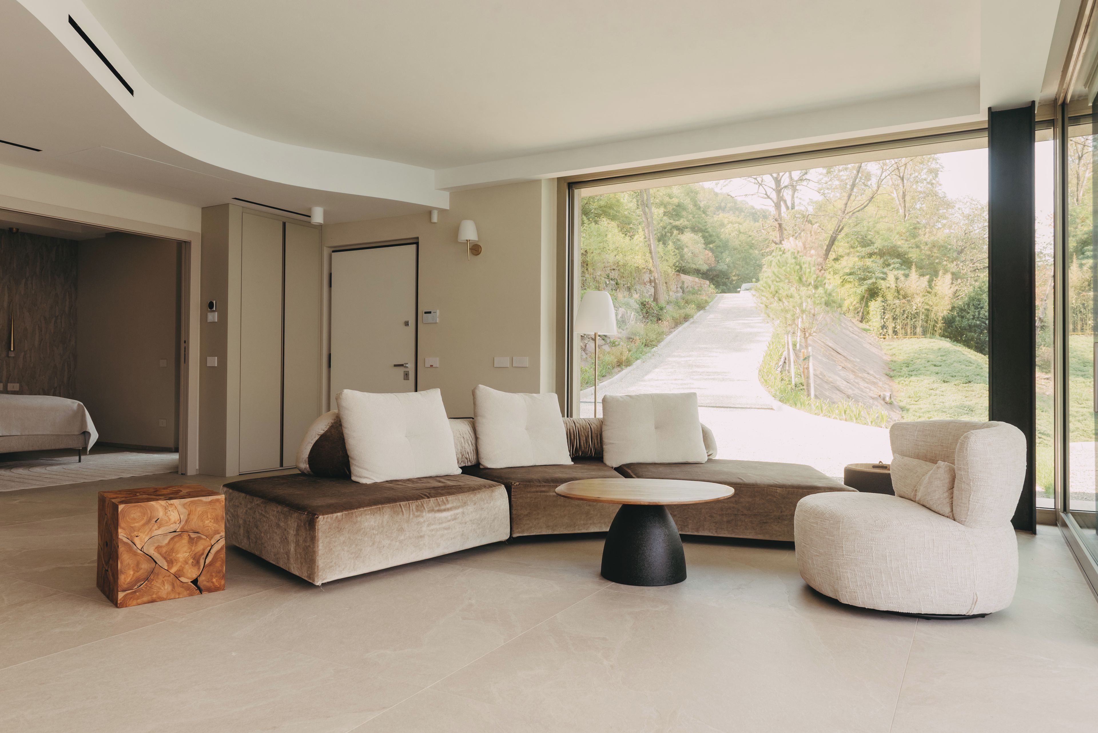 Una sala de estar moderna con un sofá modular, una mesa de centro redonda y vista a un área boscosa a través de amplios ventanales.