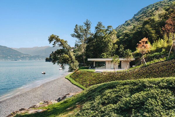 A lakeside property with a pebble beach, a small house with a patio, and lush greenery. - Villa Piccola Anna (Luino)