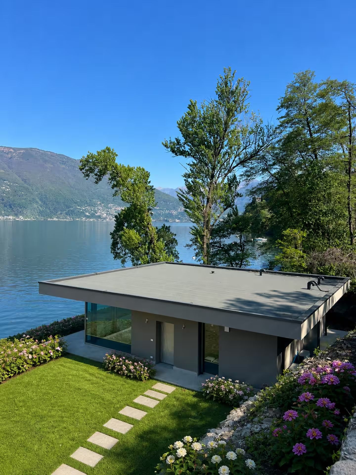 A modern house with a flat roof, large windows, and a view of a lake and mountains.