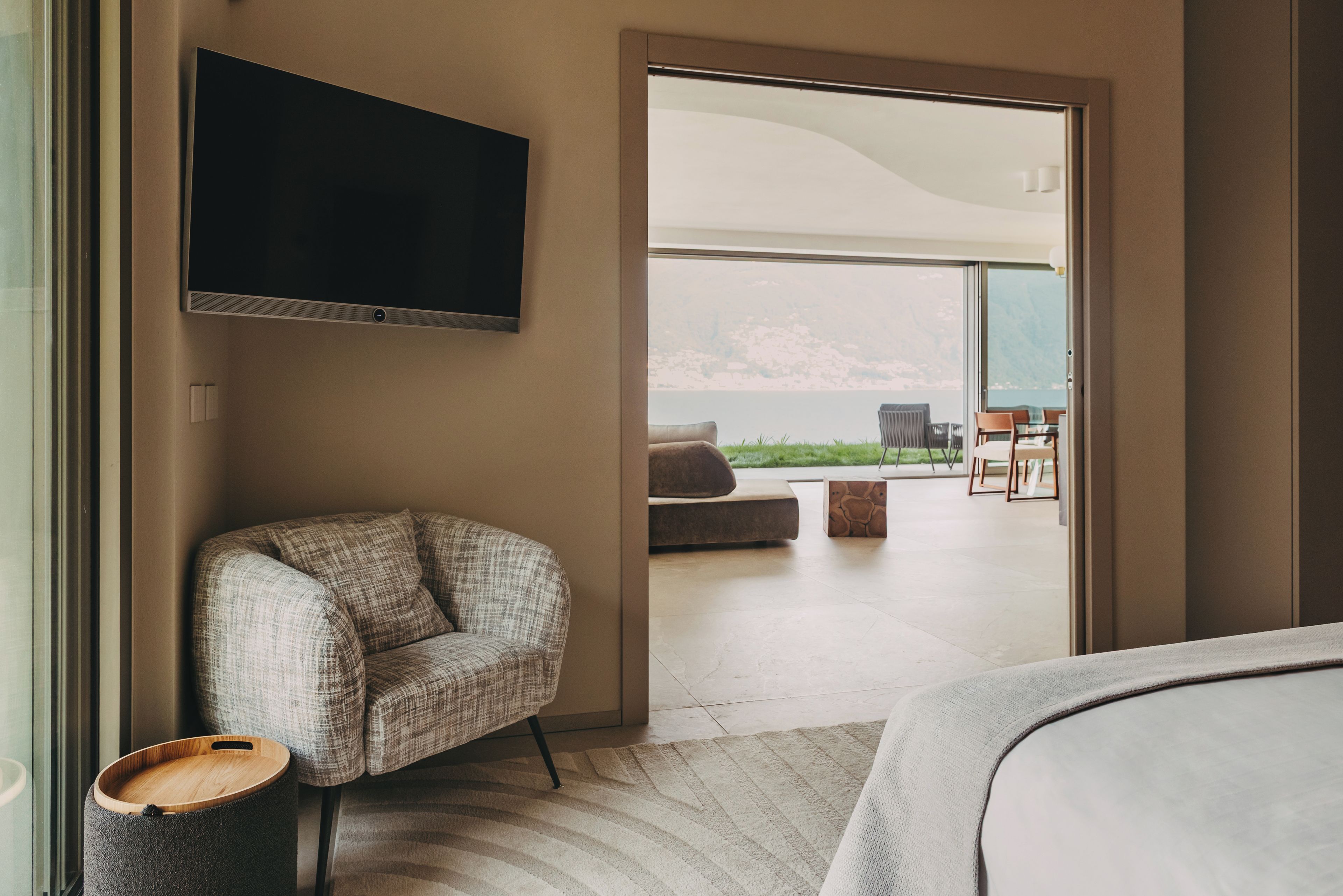 A modern hotel room with a flat-screen TV mounted on the wall, a comfortable sofa, a wooden side table, and a large sliding glass door leading to an outdoor seating area with a view of the mountains.