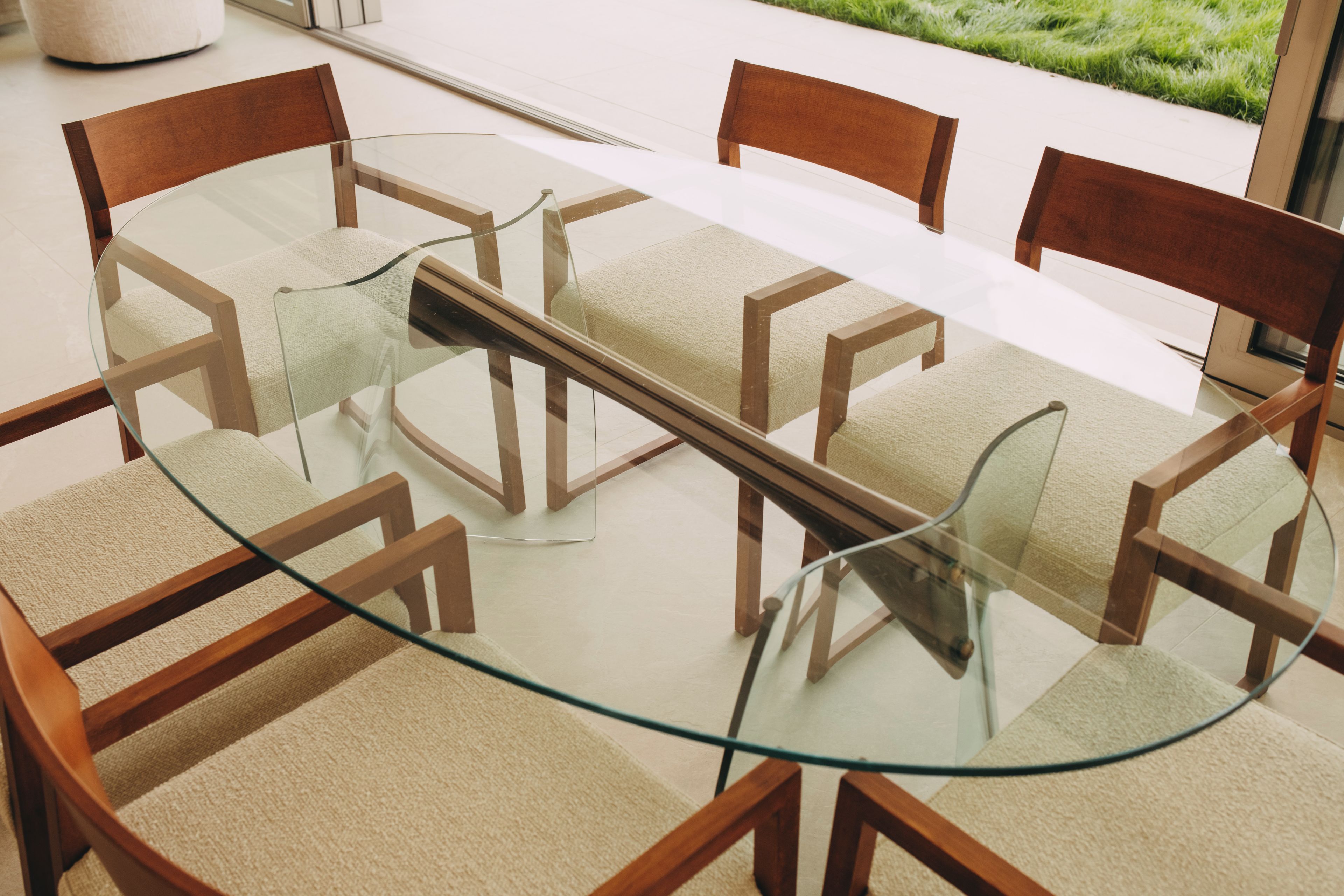 A round glass table with wooden chairs, a white floor, and a view of greenery outside.
