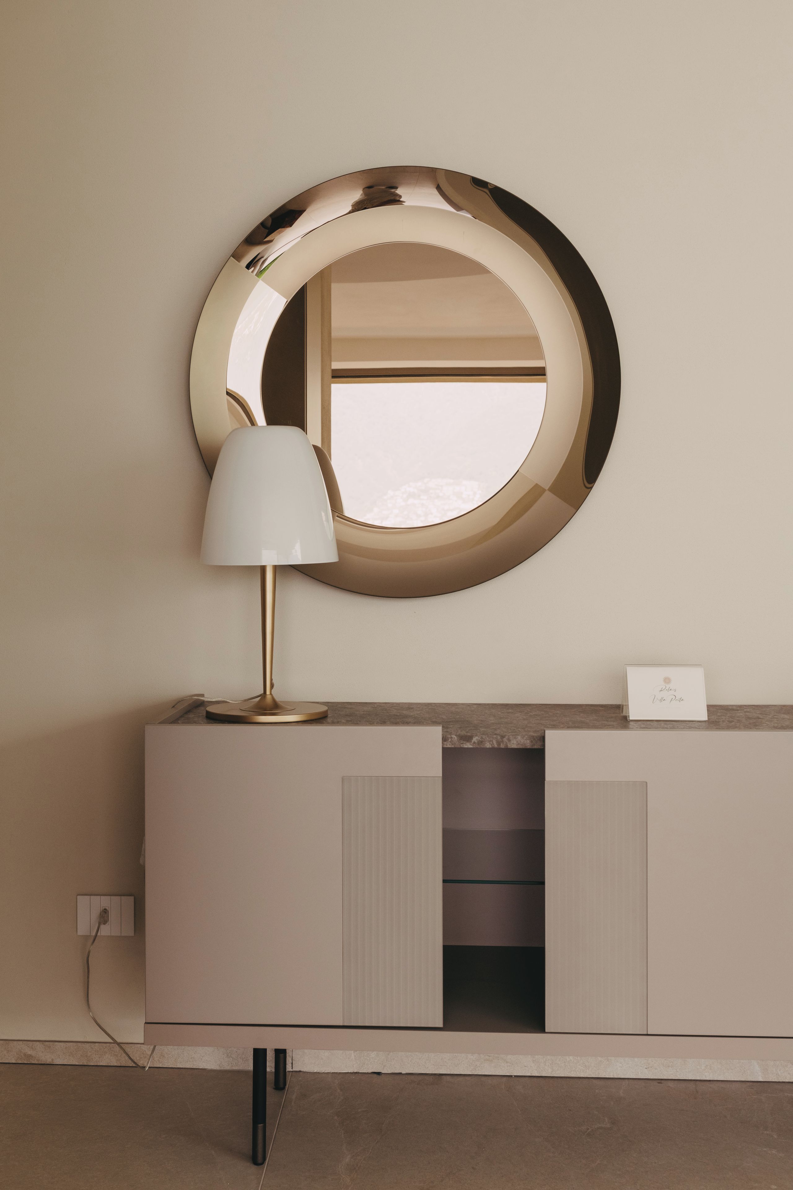 Ein moderner Waschtisch mit rundem Spiegel, einer weißen Lampe und einer kleinen Karte auf der Arbeitsfläche.