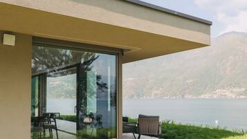 Un bâtiment moderne doté d’une porte en verre qui donne sur un espace extérieur aménagé avec une table et des chaises, offrant une vue sur un lac et des montagnes.