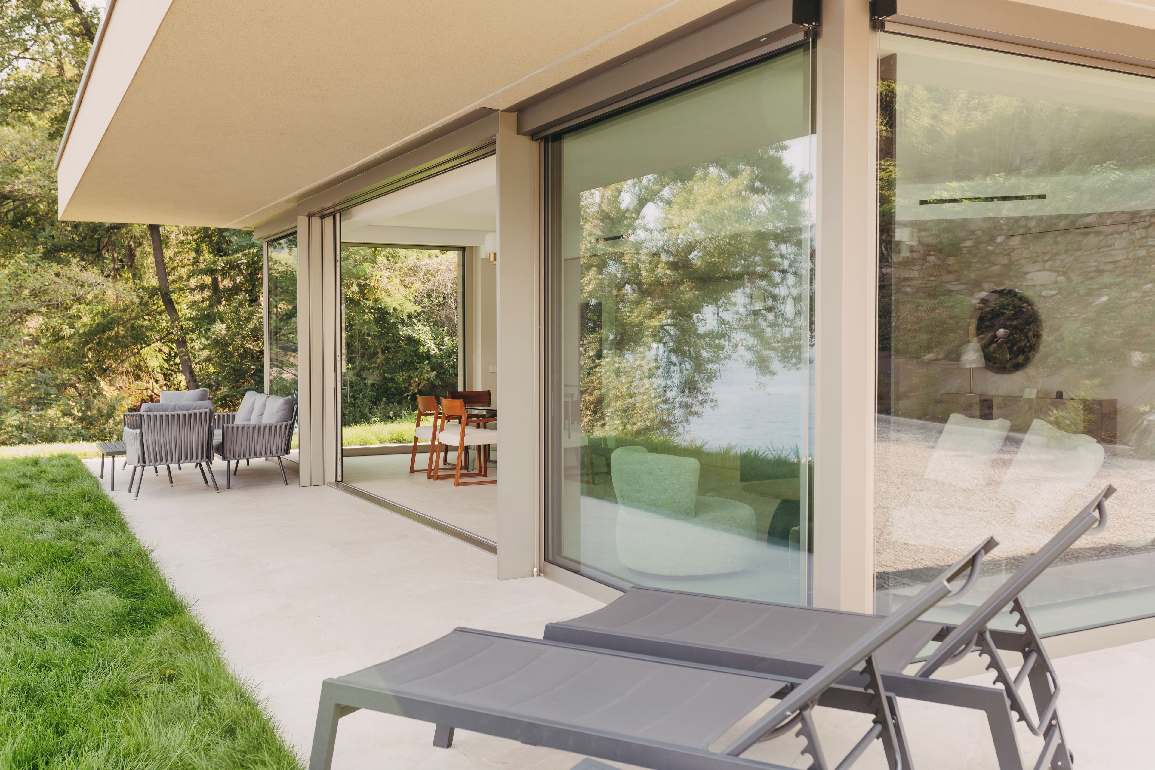 A modern outdoor patio with a glass enclosure, a dining area with chairs and a table, and a lounge area with chairs.