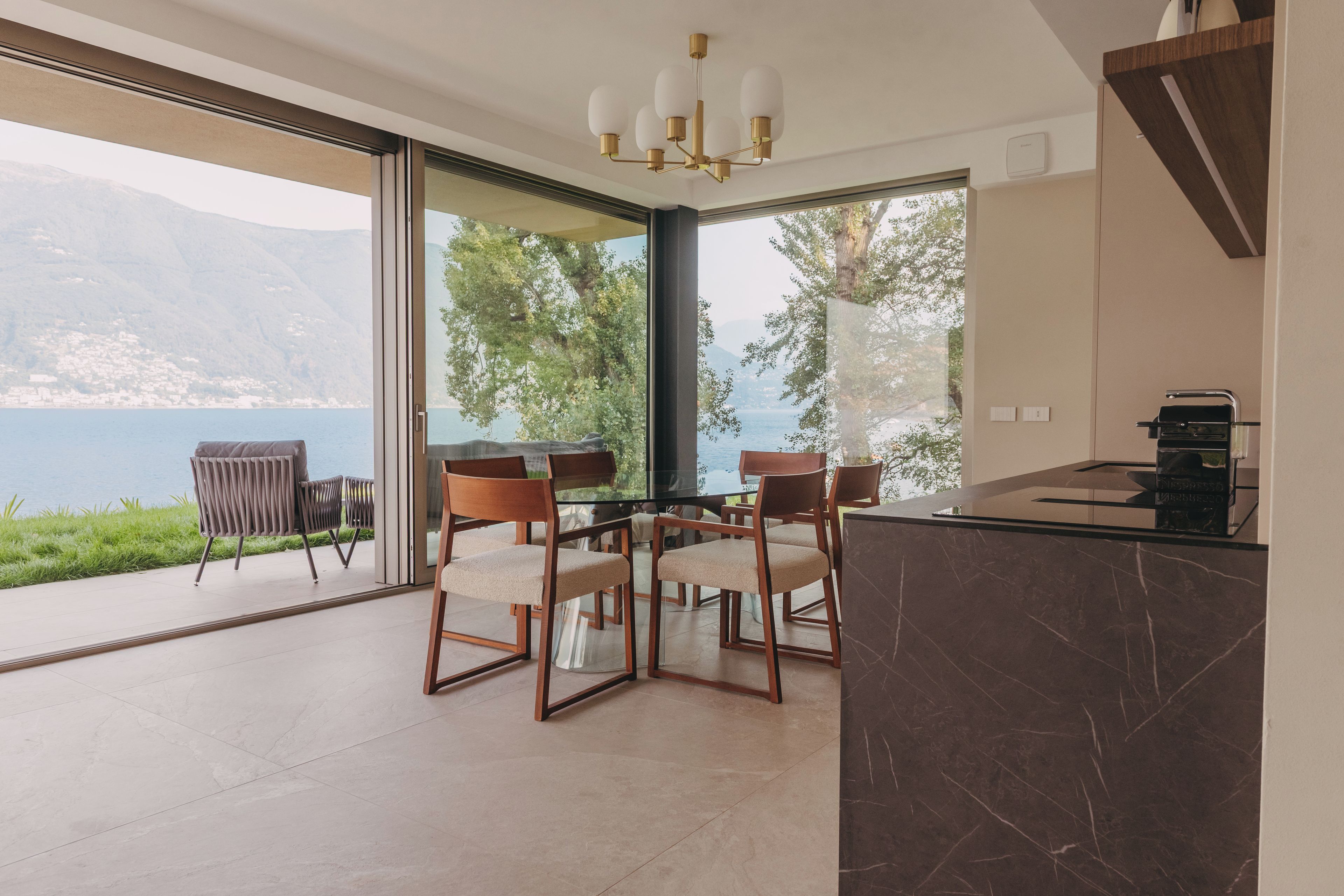 Un comedor moderno con una mesa y sillas de vidrio, una encimera de mármol y vistas a un lago y montañas.