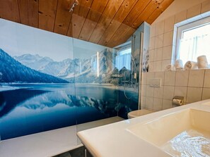 A bathroom with a large mirror reflecting a mountain landscape, a bathtub, and a window with towels on the sill. - Cabin in Annaberg With Swimming Pool (Annaberg-Lungoetz)