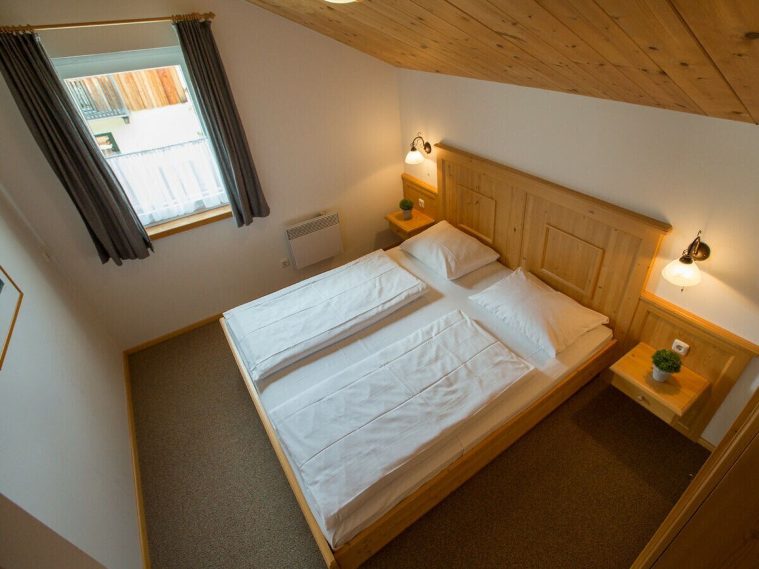 Una habitación con cama de madera, dos mesitas de noche, una ventana con cortinas y un aplique de pared.