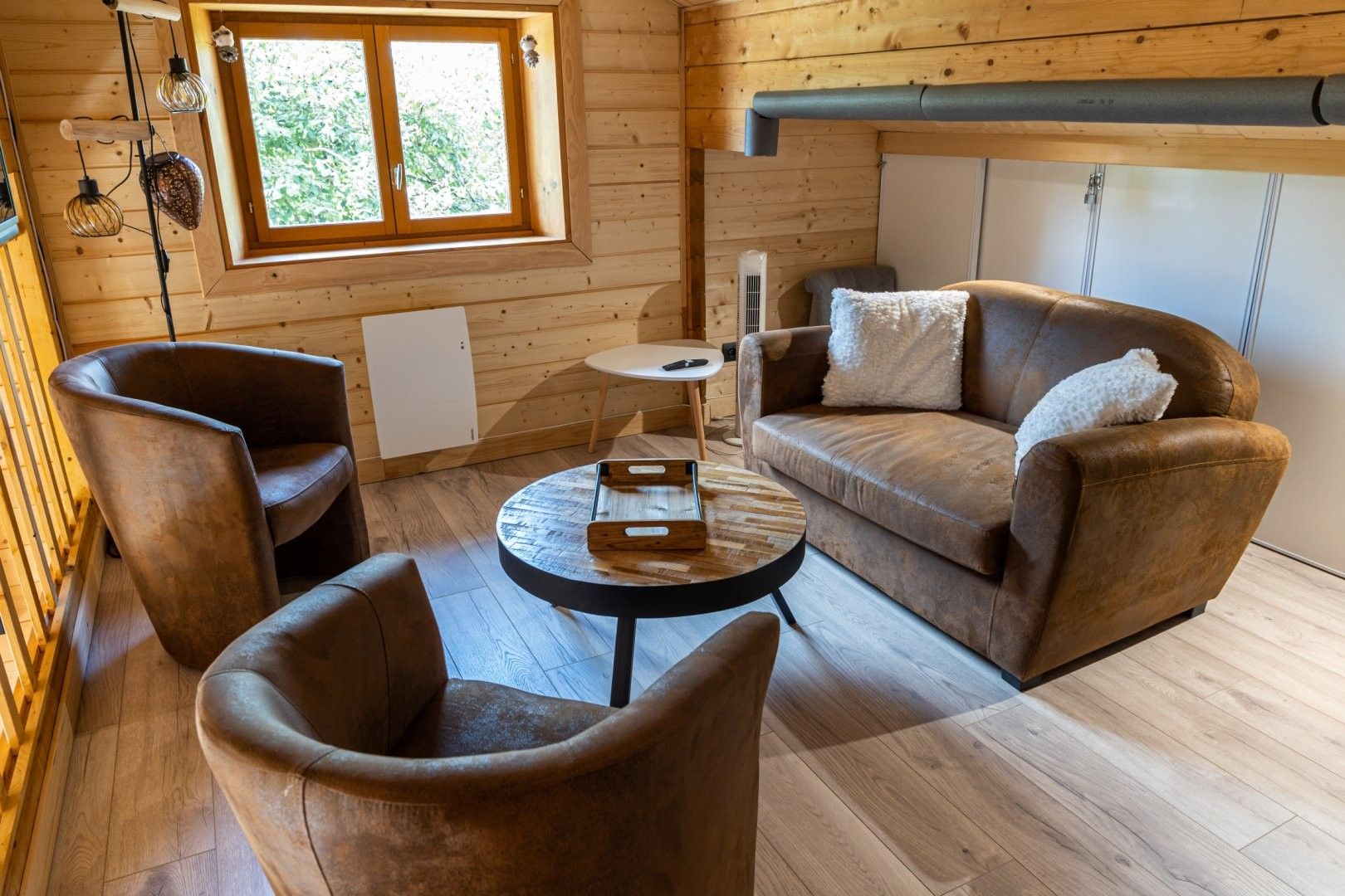 A cozy room with a wooden interior, a sofa, two armchairs, a small round table, and a window with a view of greenery.