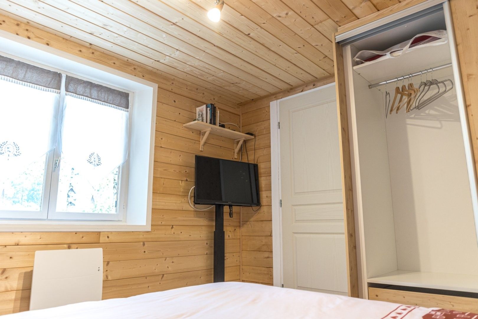 A bedroom with a bed, a television mounted on the wall, a window with curtains, and a wardrobe with hanging clothes.
