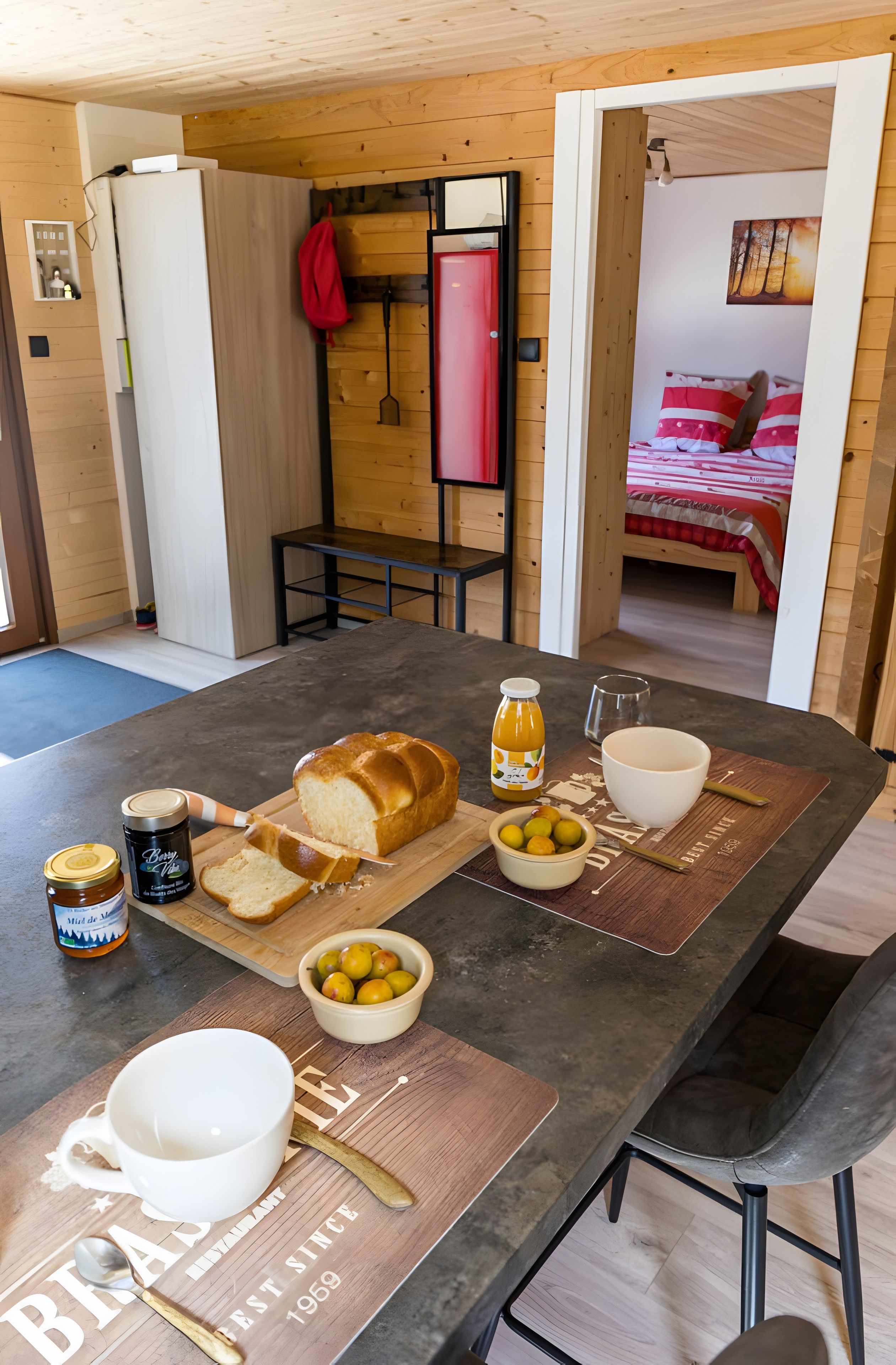 Spisebord med brød, frugt og tilbehør, et tekøkken med vask og et soveværelse med seng og et billede.