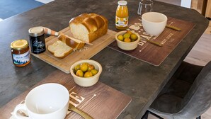 Une table à manger avec du pain, des fruits et des condiments, un coin cuisine avec un évier, et une chambre avec un lit et un tableau.