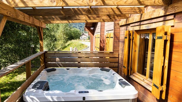 Une terrasse en bois avec un jacuzzi, des rampes en bois et une vue sur une maison au loin.
