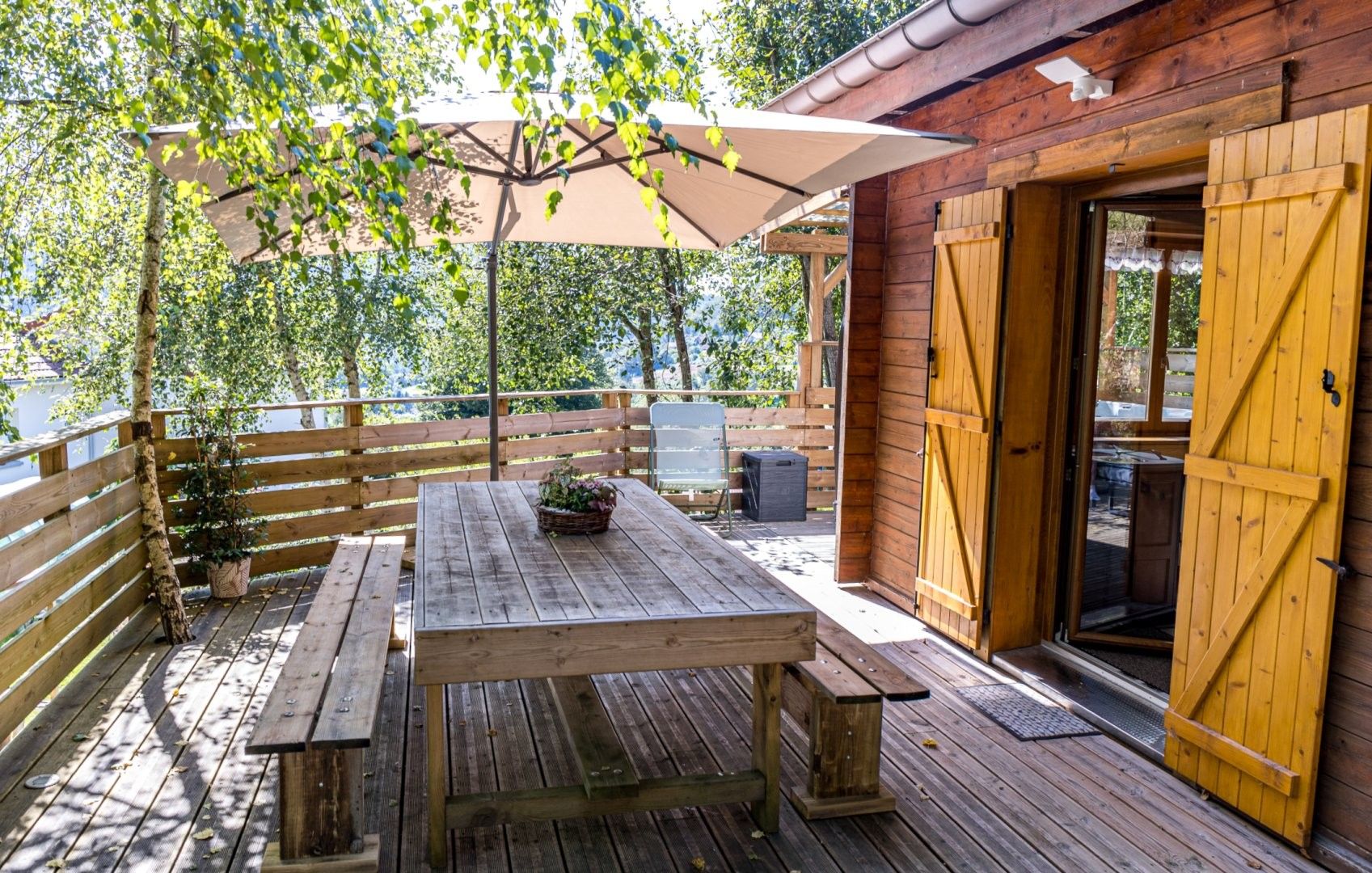 En udendørs træterrasse med et picnicbord og bænke, et træhegn og en bygning med en trædør.