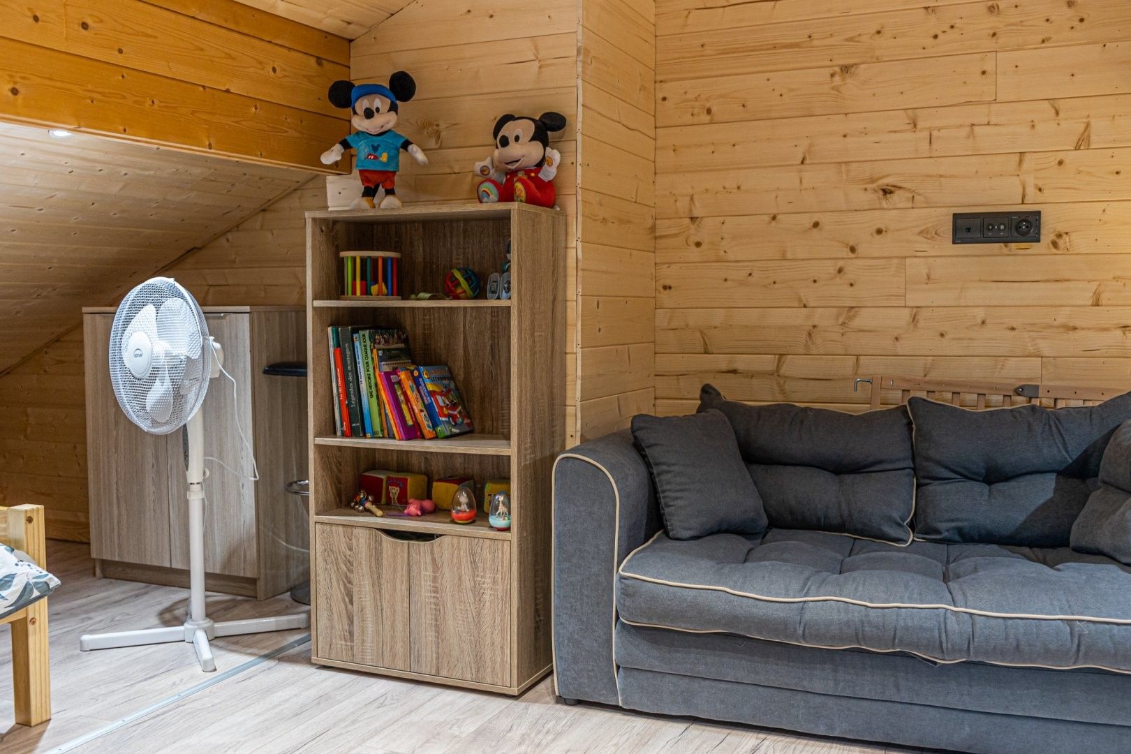 Una habitación acogedora con una estantería de madera, un sofá gris y dos peluches encima.