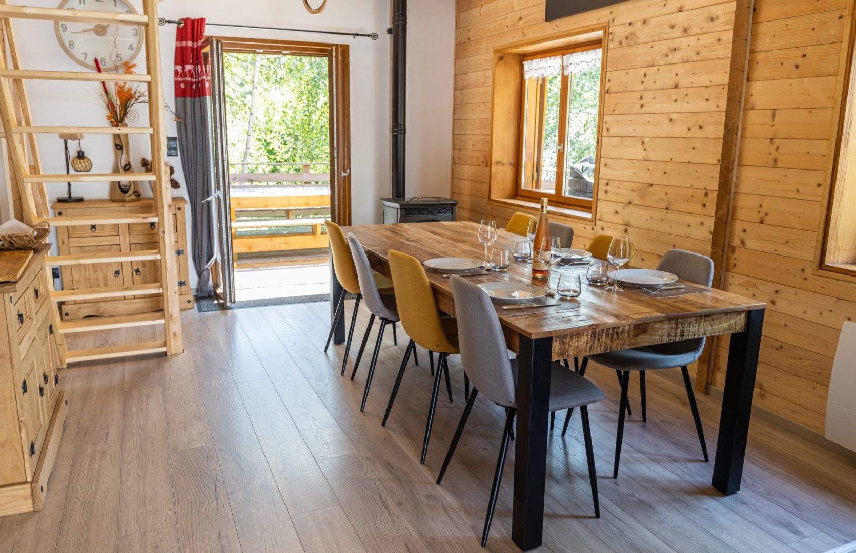 Interior de madeira com mesa de jantar posta para quatro, um armário de madeira com gavetas e janela com vista para o verde.