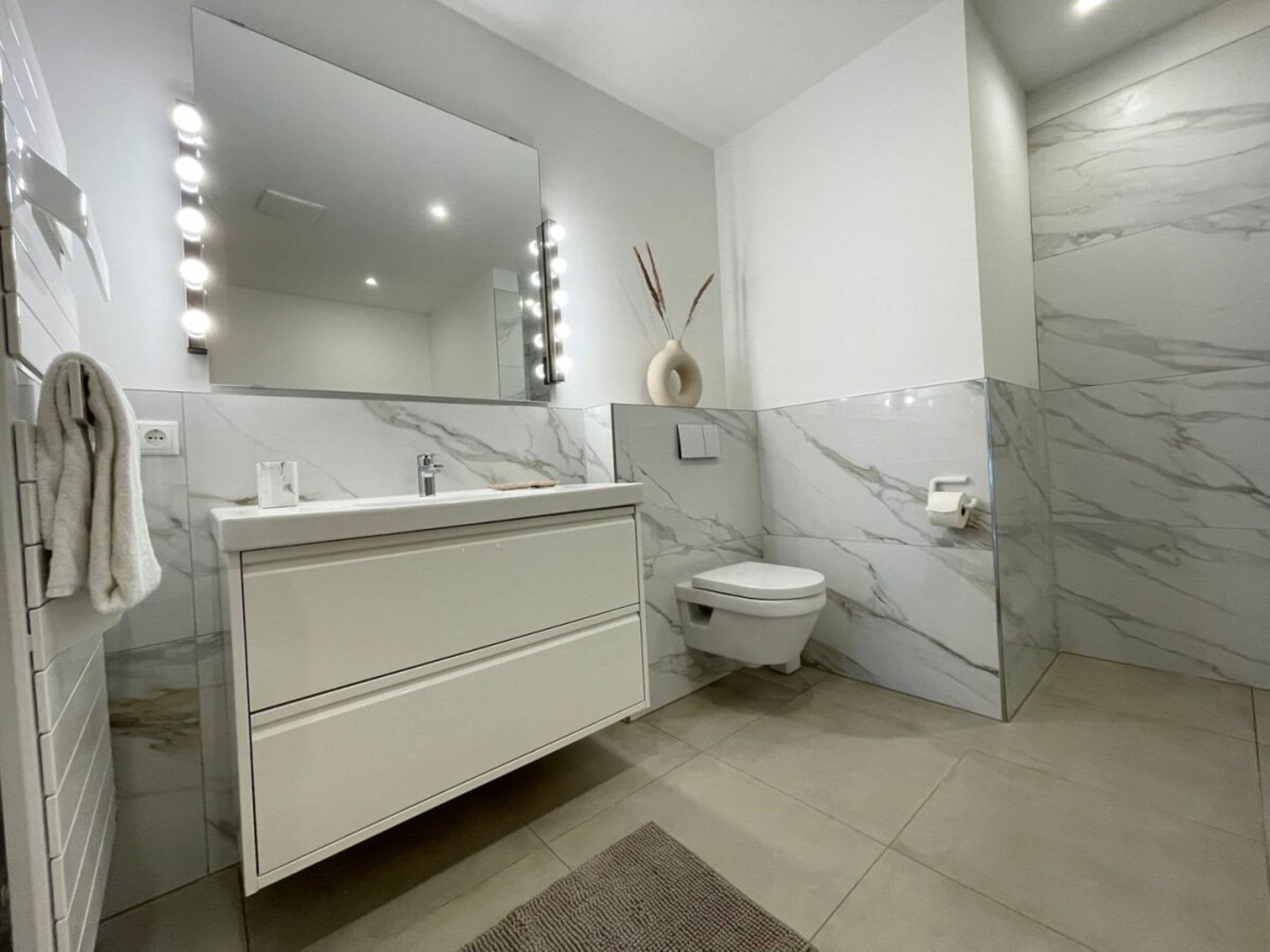 A modern bathroom with a marble vanity, a toilet, and a large mirror with lights.