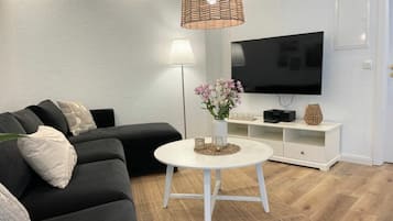 Una sala moderna con un sofá negro, una mesa de centro blanca, un televisor de pantalla plana y una lámpara colgante de fibras naturales.