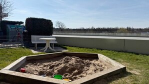 Zona arenosa para jugar con un banco y una mesa, rodeada por una valla y un seto.