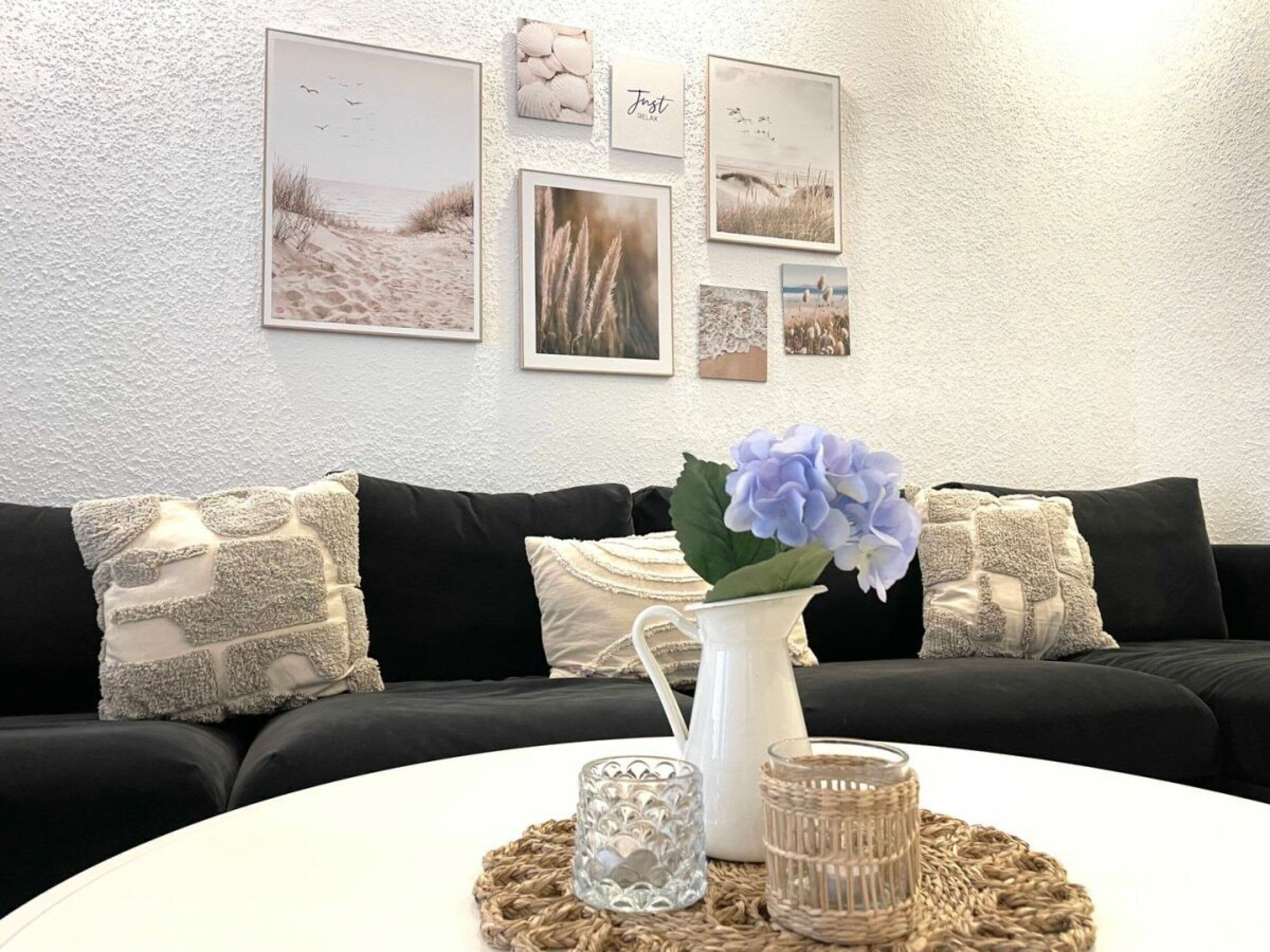 A living room with a black sofa, decorative pillows, a white vase with flowers, and wall art.
