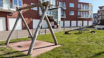 Un parque infantil con columpios y un tobogán, rodeado de césped y un edificio de ladrillo con ventanas y balcones.