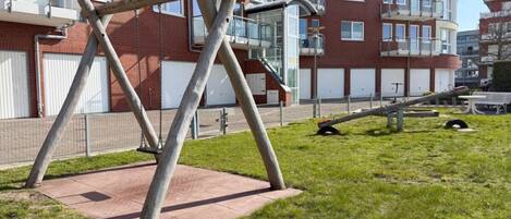 Un parque infantil con columpios y un tobogán, rodeado de césped y un edificio de ladrillo con ventanas y balcones.