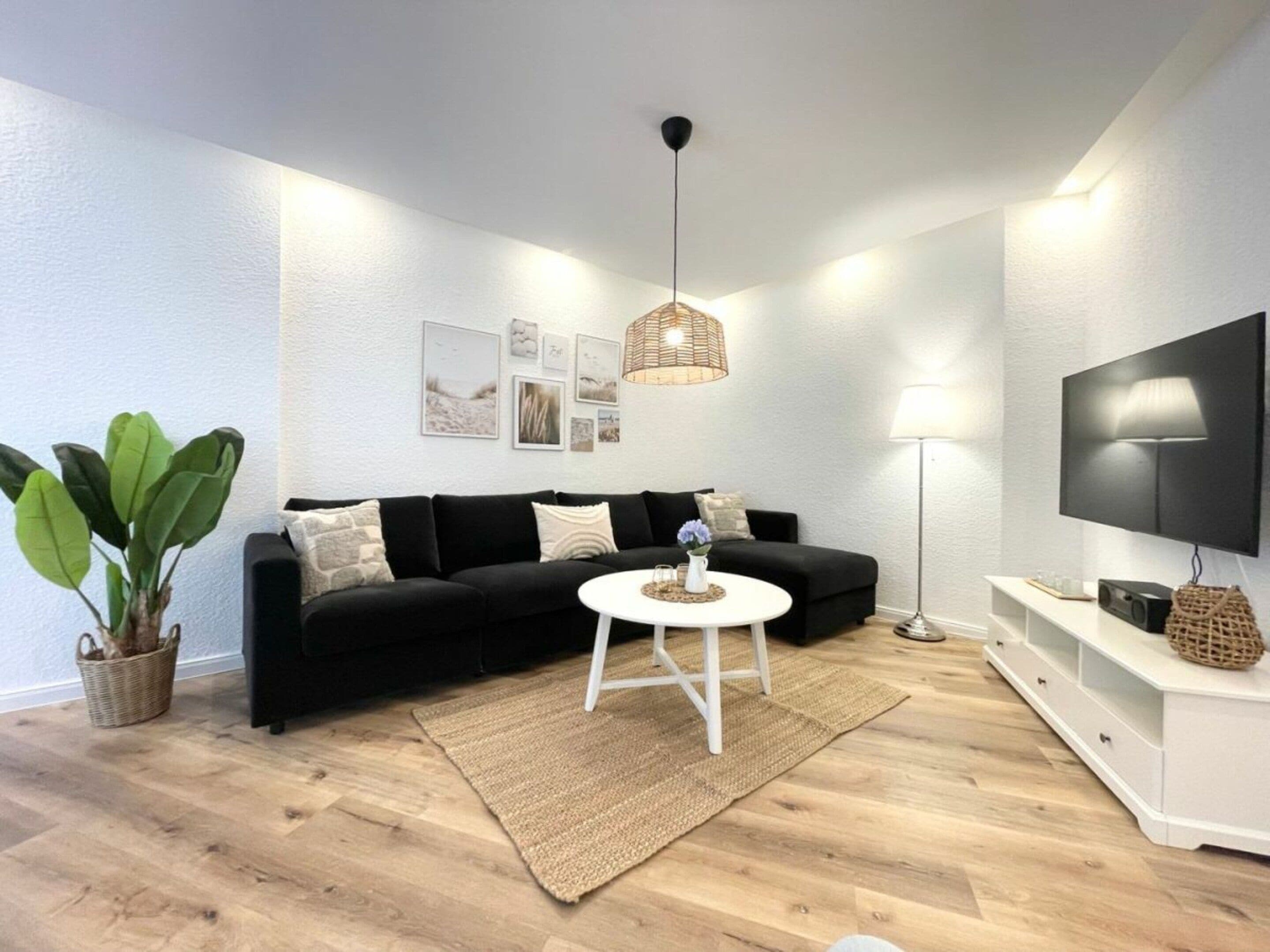 Una sala de estar moderna con un sofá negro, una mesa de centro blanca, una lámpara de pie, un televisor en la pared y una planta.