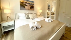 Una cama bien hecha con dos toallas enrolladas, una lámpara de noche, una mesita de noche, un espejo y un cuadro de una playa en la pared.