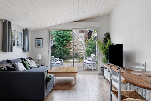 Una sala de estar moderna con un sofá gris, una mesa de centro de madera, un televisor y un suelo de vidrio corredera que da a un patio exterior con sillas y una mesa blancos.